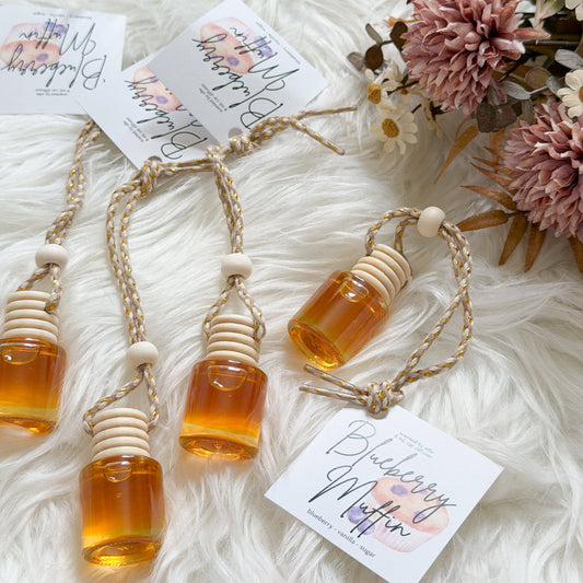Three small glass honey bottles with wooden dipper tops and braided string, arranged on a white faux-fur surface with floral props and printed tags reading Blueberry Muffin.