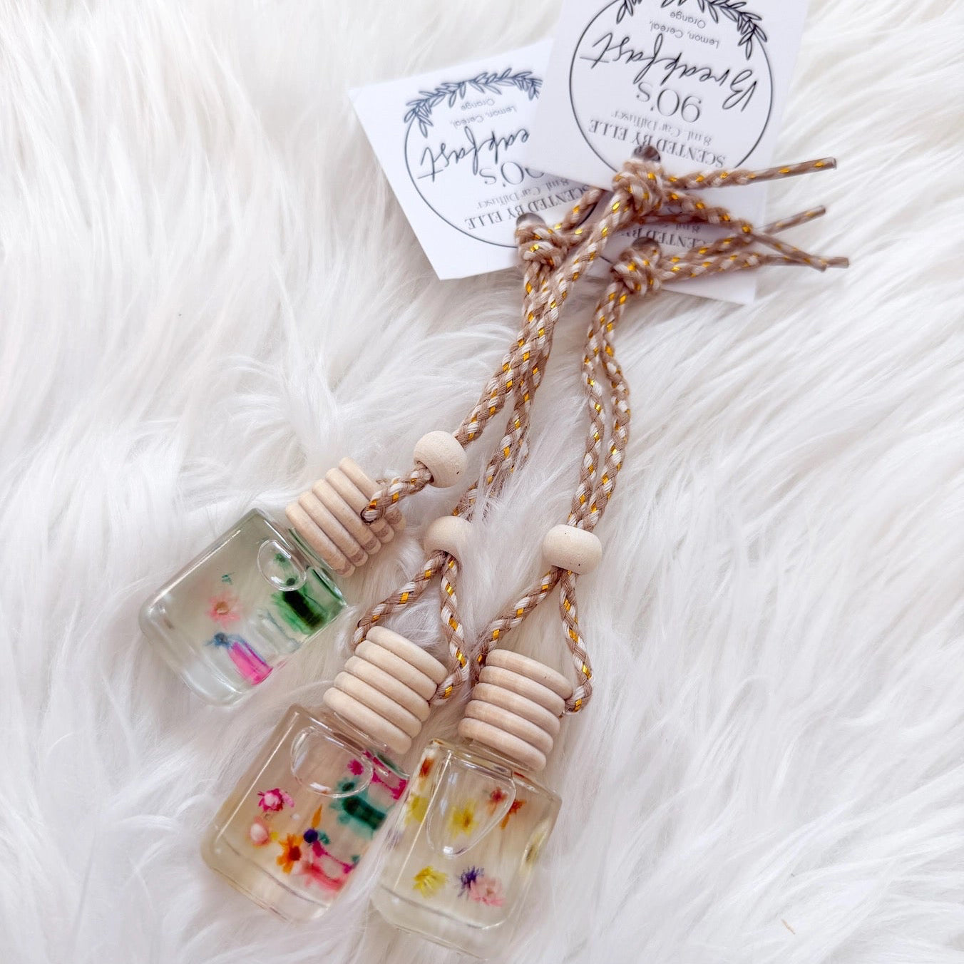 Three small glass bottles with wooden tops on a white textured surface, featuring a brand tag.
