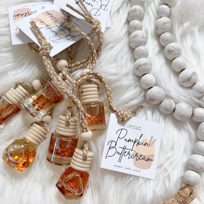 A styled flat-lay of resin glass diffusers with amber-colored liquid, tied with jute twine and attached to white cards reading 'Pumpkin Buttercream' and 'Pumpkin Spice'. A white faux fur rug, a white beaded garland, and a beige tassel are arranged around them.