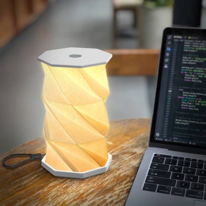 A modern geometric folding table lamp with a warm yellow glow, sitting on a wooden desk next to an open laptop.