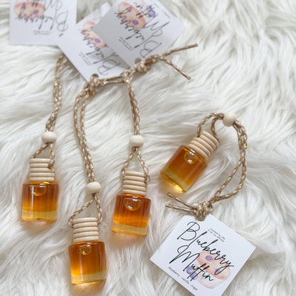 Three amber glass bottle-shaped perfume oil vials with beige cotton wicks and braided cords, displayed on a white faux fur surface with printed tags.