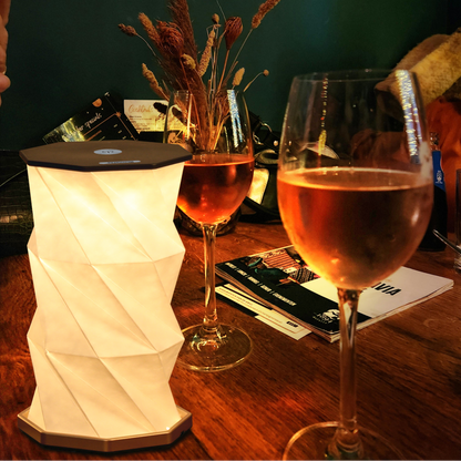 A modern white faceted geometric table lamp glowing warm white on a wooden table, next to two wine glasses filled with rosé wine and a vase of dried grasses against a dark green wall.