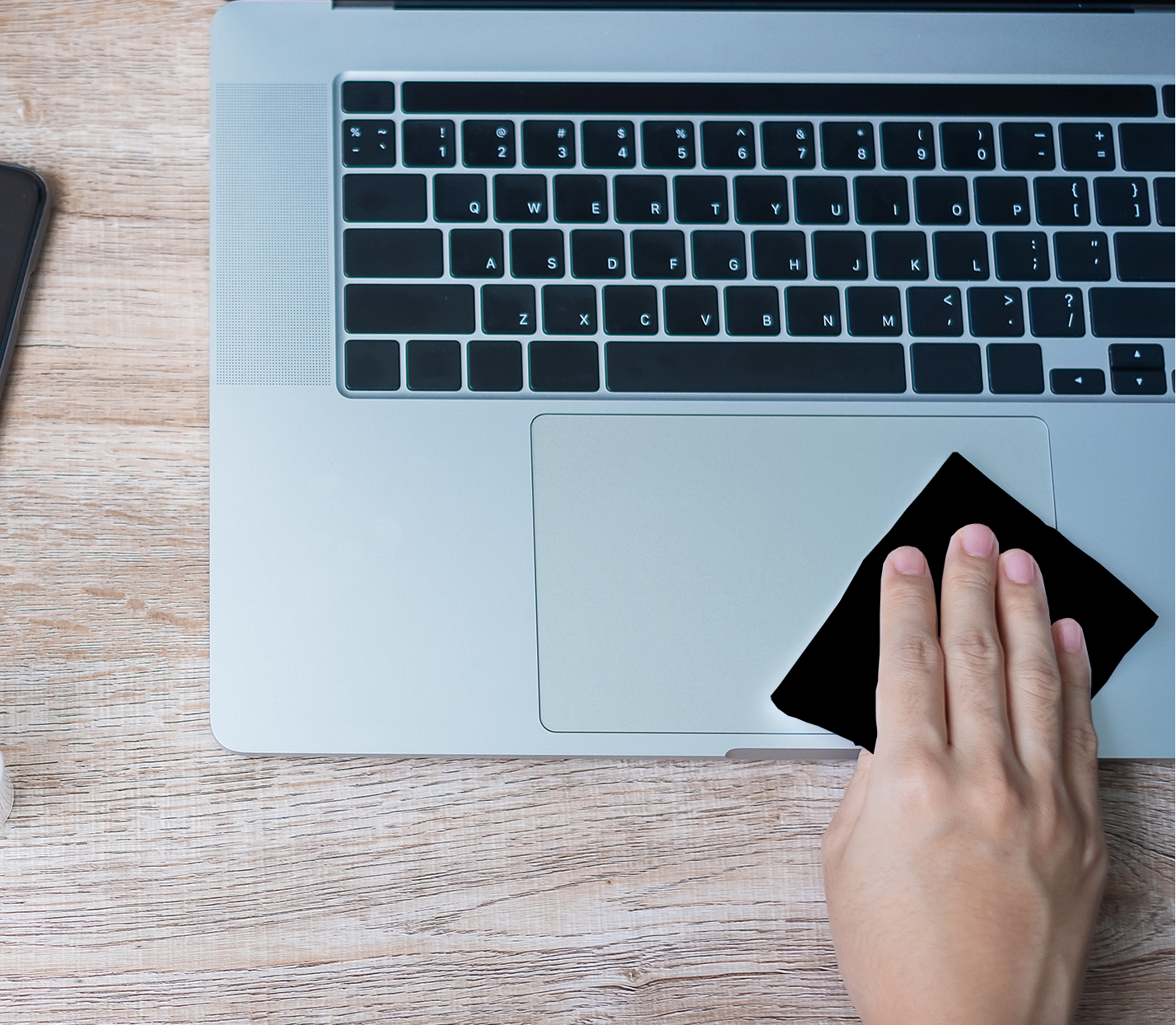 Person cleaning a laptop keyboard with a black cloth on a wooden desk