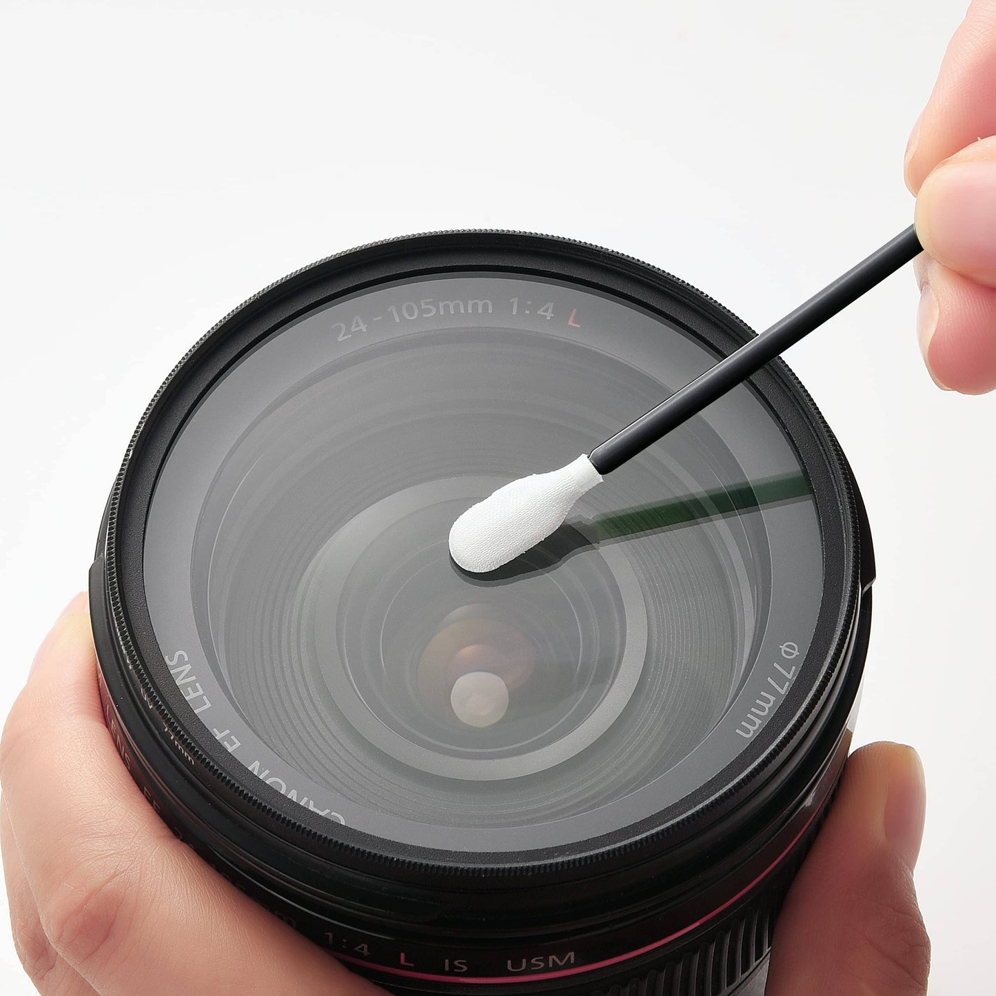 A close-up of a camera lens being cleaned with a cotton swab held by a hand, against a clean white background.