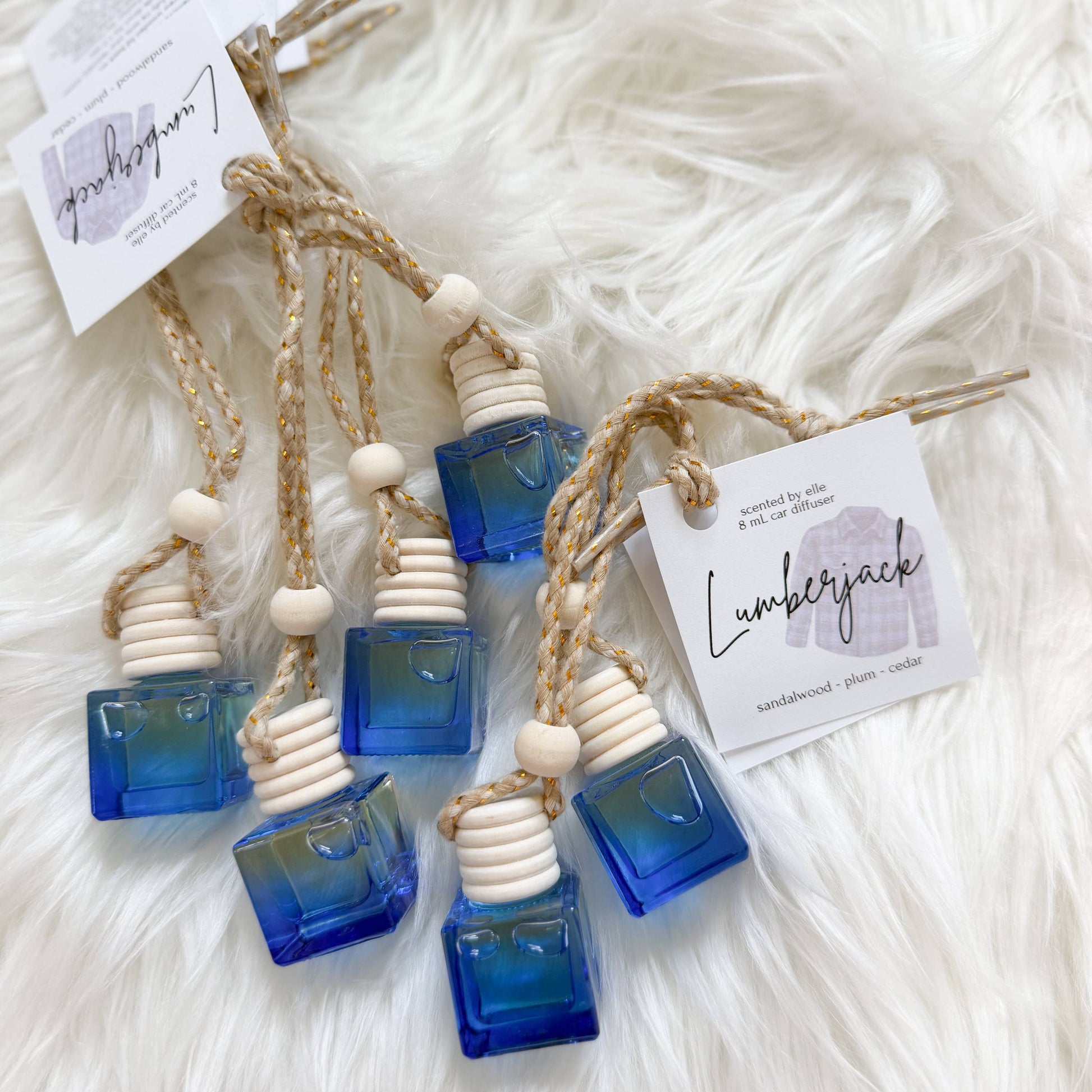 Five amber glass bottle reed diffusers with blue gradient liquid, natural wood beads, and jute cords, each with a branded paper tag, displayed on a white faux fur background.
