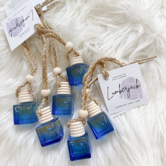 Five amber glass bottle reed diffusers with blue gradient liquid, natural wood beads, and jute cords, each with a branded paper tag, displayed on a white faux fur background.