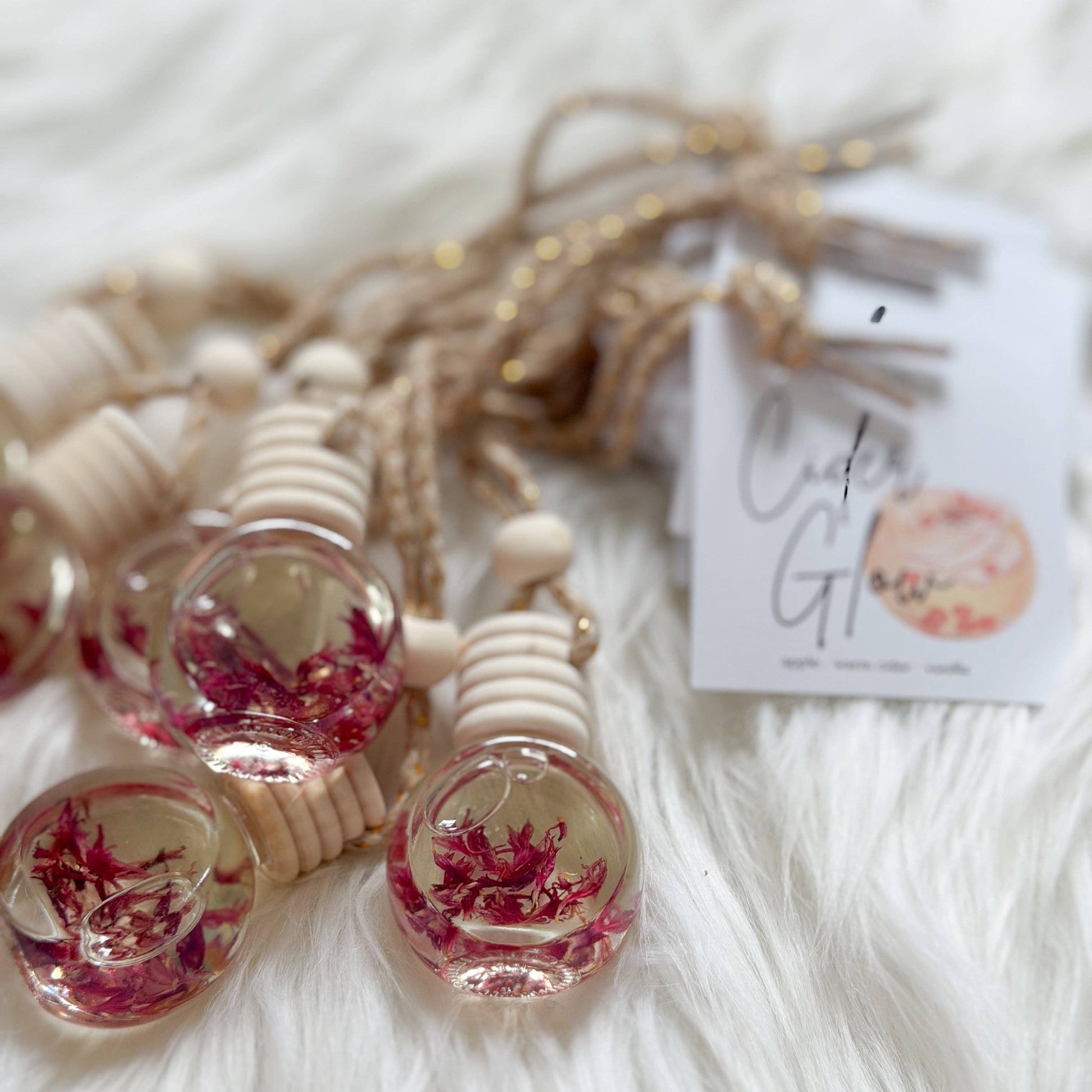 Several small glass vial charms filled with pink dried flowers, attached to beige cord and wooden beads, arranged on a white faux-fur surface with a small printed tag.