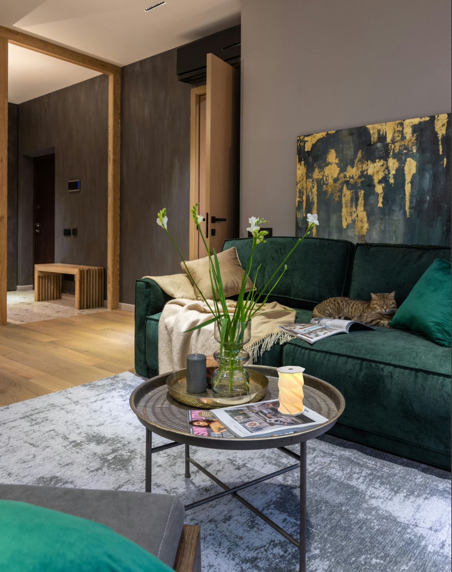 A modern living room featuring a plush dark green velvet sofa with a cat resting on it, a round metal coffee table in the foreground, and a large abstract wall art piece with gold and black tones above the couch.