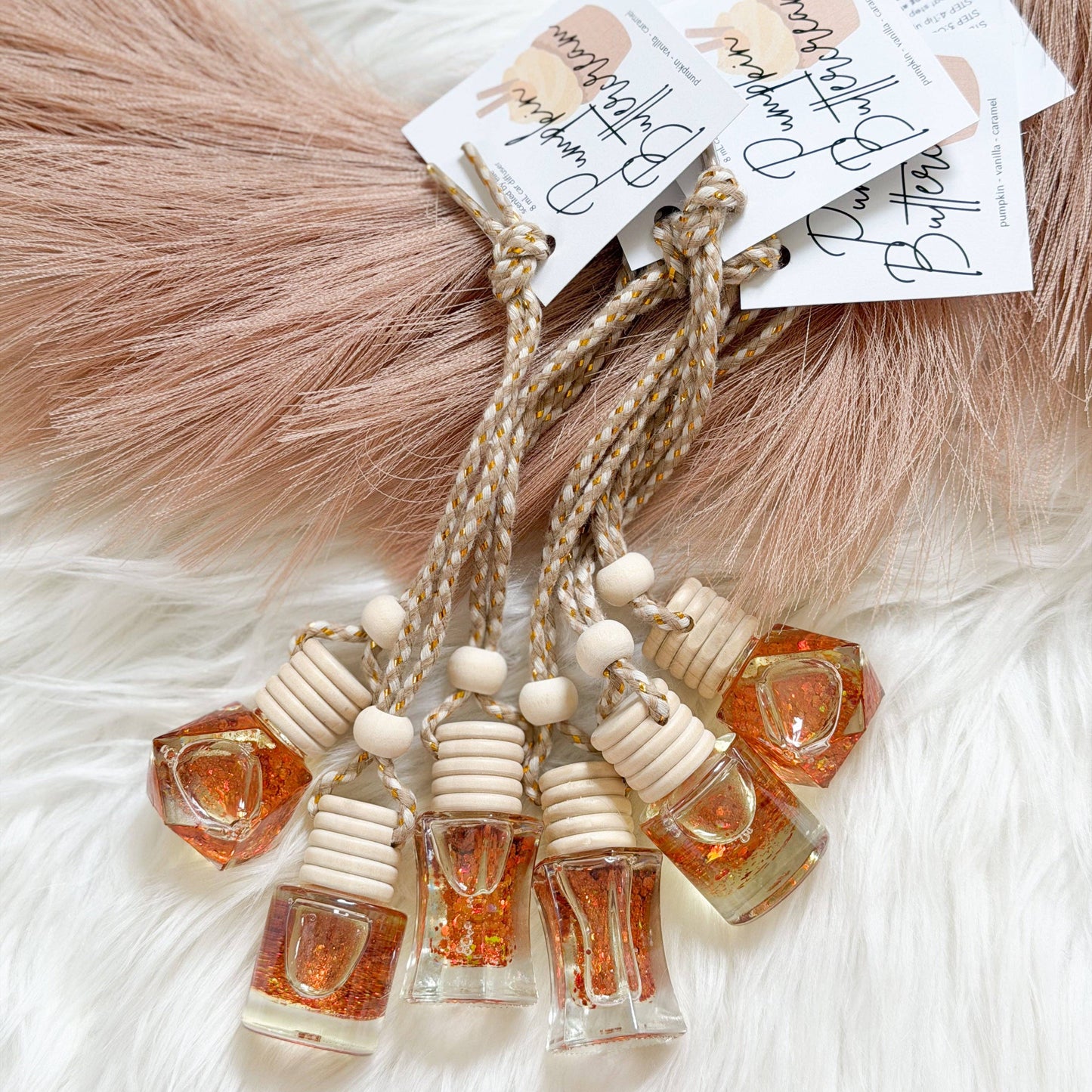 Four amber glass bottle diffusers with white wooden diffuser tops and beige braided cords, each attached to a white paper tag, arranged on a white faux fur surface with a dusty pink faux fur accent.