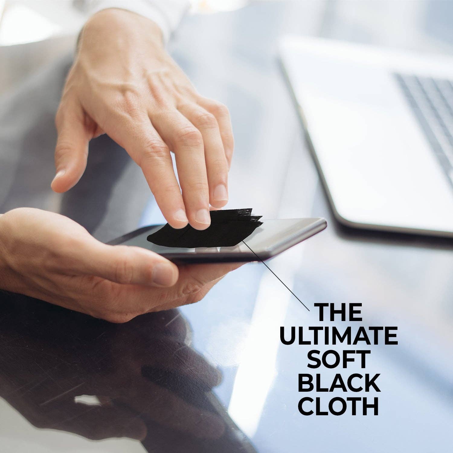Person cleaning a smartphone screen with a soft black cloth, blurred background with laptop.