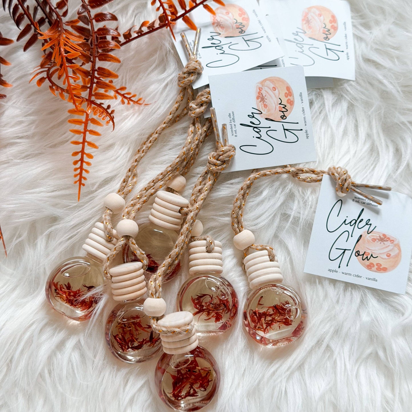Three glass bottle car air fresheners filled with dried red botanicals, attached to natural wood and jute cords, displayed on a white faux fur background with decorative ferns and branded tags reading 'Cider Glow'.