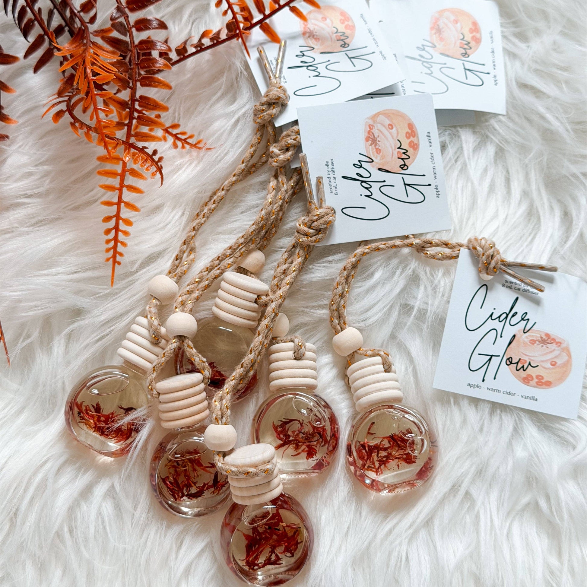 Three glass bottle car air fresheners filled with dried red botanicals, attached to natural wood and jute cords, displayed on a white faux fur background with decorative ferns and branded tags reading 'Cider Glow'.