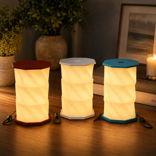 Three compact folding lanterns with ribbed cylindrical bodies are arranged on a wooden table in a cozy indoor setting. The left lantern has a dark red lid, the center one has a light gray lid, and the right one has a teal-blue lid.