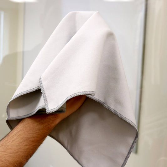 A close-up of a hand holding a light gray microfiber cloth with gray stitched edges, set against a softly blurred indoor background.