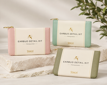 Three rectangular earbud cleaning pouches labeled 'EARBUD DETAIL KIT' by PINCH are arranged on stacked stone slabs. The pouches have cream centers with pink, mint green, and sage green sides, and small gold zipper pulls.