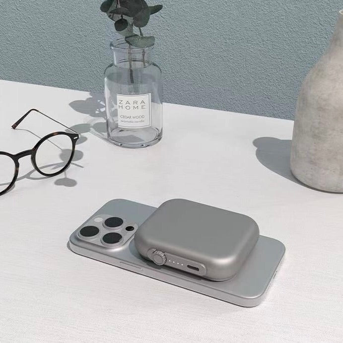A gray rectangular magnetic wireless charging bank holding a silver smartphone, with a matching gray portable power bank resting on top. The setup is styled on a white tabletop with a glass bottle, eyeglasses, and a ceramic vase in the background.