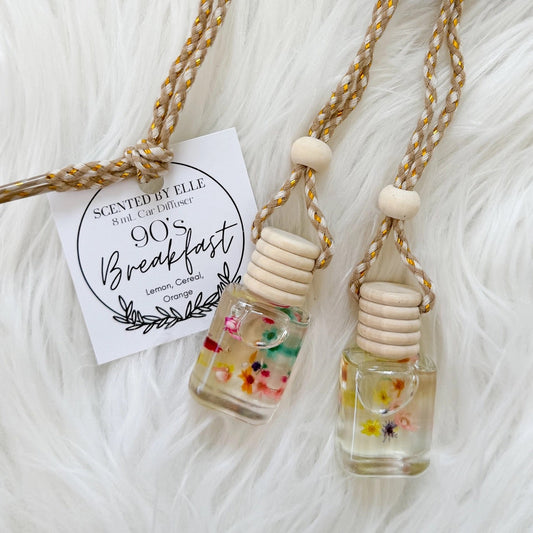 Two small glass bottles with floral designs on a white fluffy surface, featuring a '90s Breakfast' label.