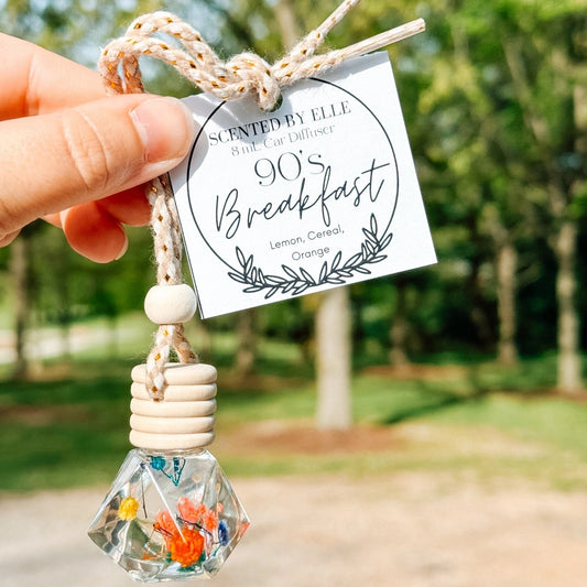 Hand holding a decorative hanging scented by Elle 90's breakfast car diffuser with a label outdoors