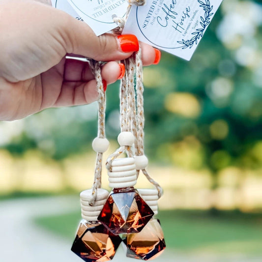 Hand holding a car air freshener with 'Scented by Elle Coffee House' tags against a blurred green background