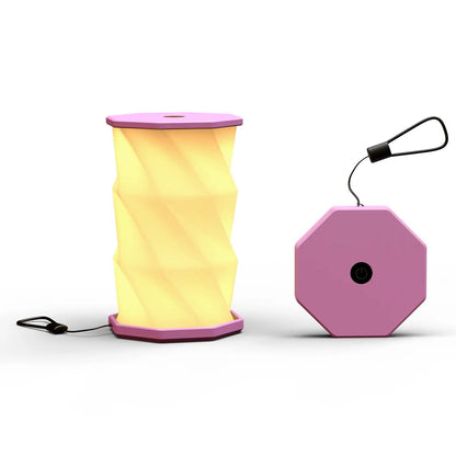 A blush folding lamp  shown on a clean white background. The lamp is unfolded and illuminated with a geometric, faceted design in warm yellow.