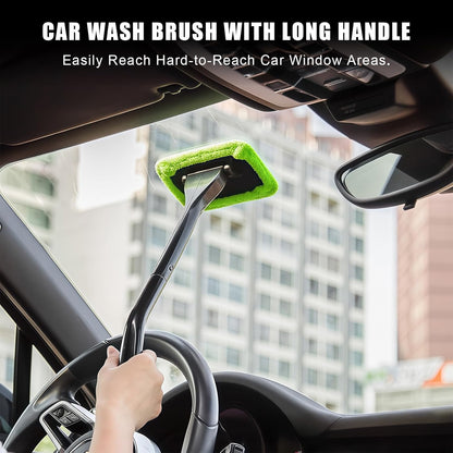 A long-handled car wash brush with a bright green microfiber head is being used inside a car to clean the windshield. A hand is visible holding the handle, and the interior of the car and city buildings are in the background.