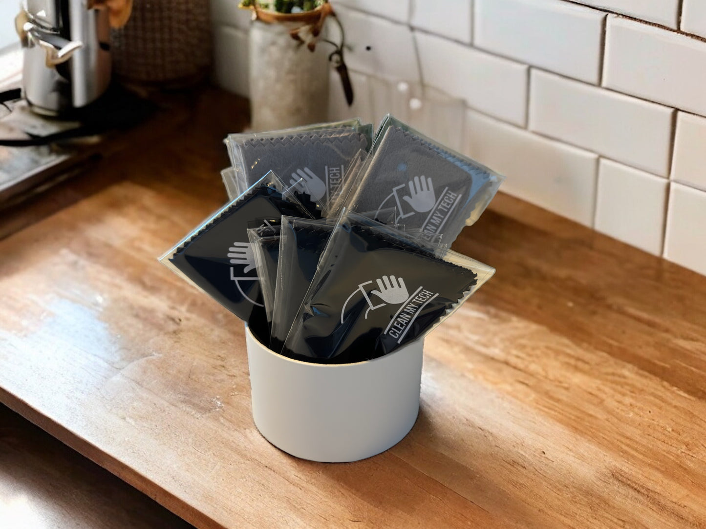 A container of individually wrapped black and grey Clean My Tech microfiber cloths sitting on a kitchen counter.