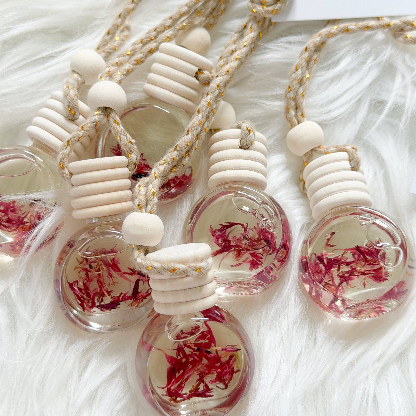 Several small clear glass pendant bottles with cream-colored wooden beads and braided rope necklaces, each containing dried pinkish-red flower petals, arranged on a white faux-fur surface.