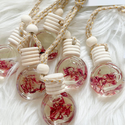 Several small clear glass pendant bottles with cream-colored wooden beads and braided rope necklaces, each containing dried pinkish-red flower petals, arranged on a white faux-fur surface.