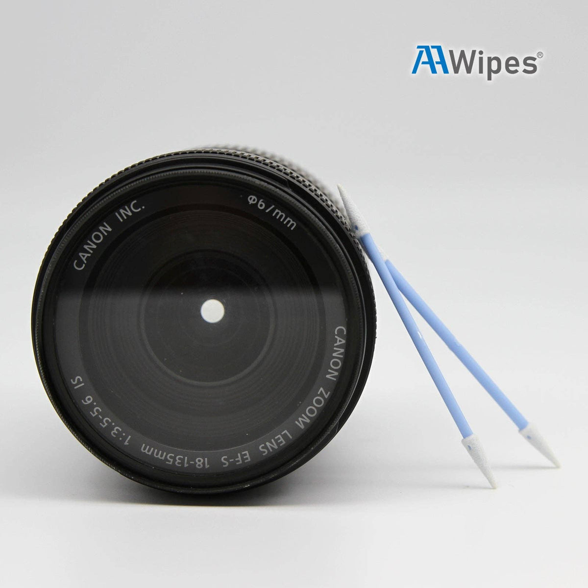 A Canon zoom lens shown on a light gray background with two blue and white cleaning wipes from AA Wipes leaning against it. The Canon brand is clearly printed on the lens.