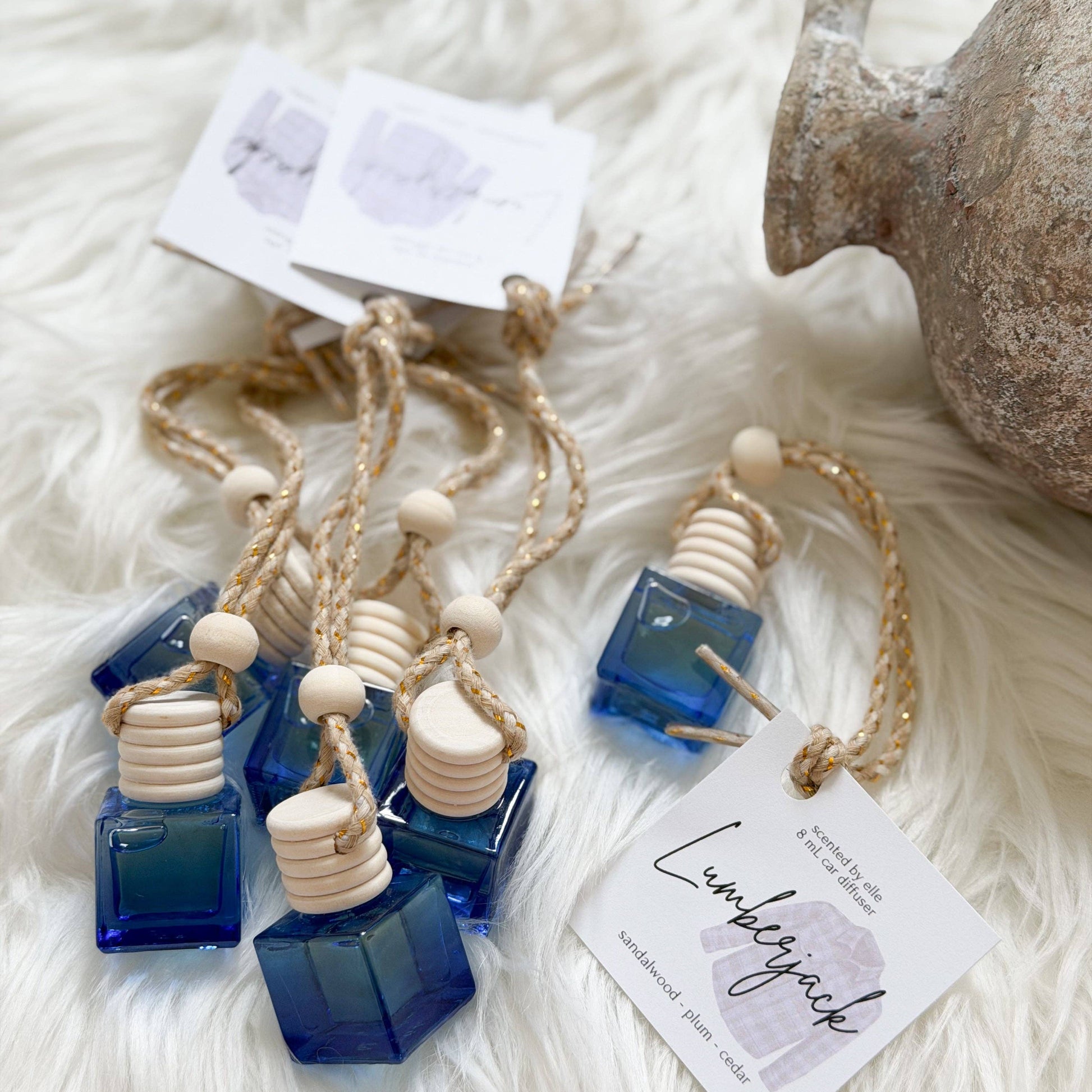 Several small blue glass bottle-shaped ornaments with natural wood beads and jute cords are arranged on a white faux-fur surface. One tag reads 'Lumberjack' and 'handcrafted by Lumberjack,' and a rustic stone jug is partially visible at the upper right.
