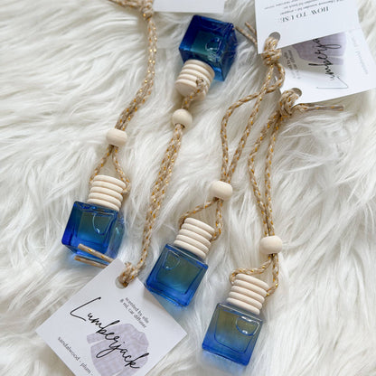 Three handmade amber glass perfume atomizer bottles with blue and green gradient glass cubes, cream-colored wooden beads, and braided jute cords, displayed on a white faux fur background with product tags.