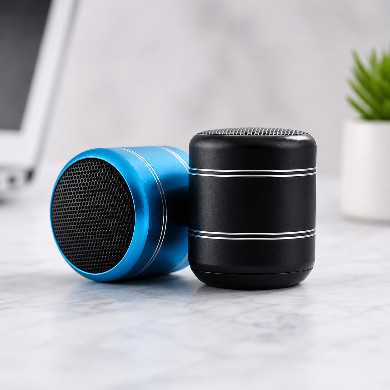 Two portable cylindrical speakers on a light marble surface, one in metallic blue and one in black, with a blurred laptop and small plant in the background.