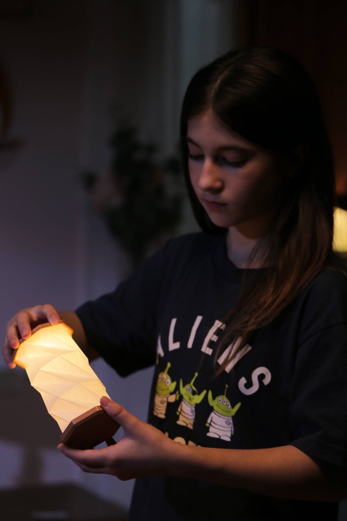 A young woman holding a glowing, illuminated paper lantern in a dimly lit indoor setting. She is wearing a dark graphic t-shirt featuring the word 'ALIENS' and three green alien characters.