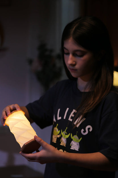 A young woman holding a glowing, illuminated paper lantern in a dimly lit indoor setting. She is wearing a dark graphic t-shirt featuring the word 'ALIENS' and three green alien characters.
