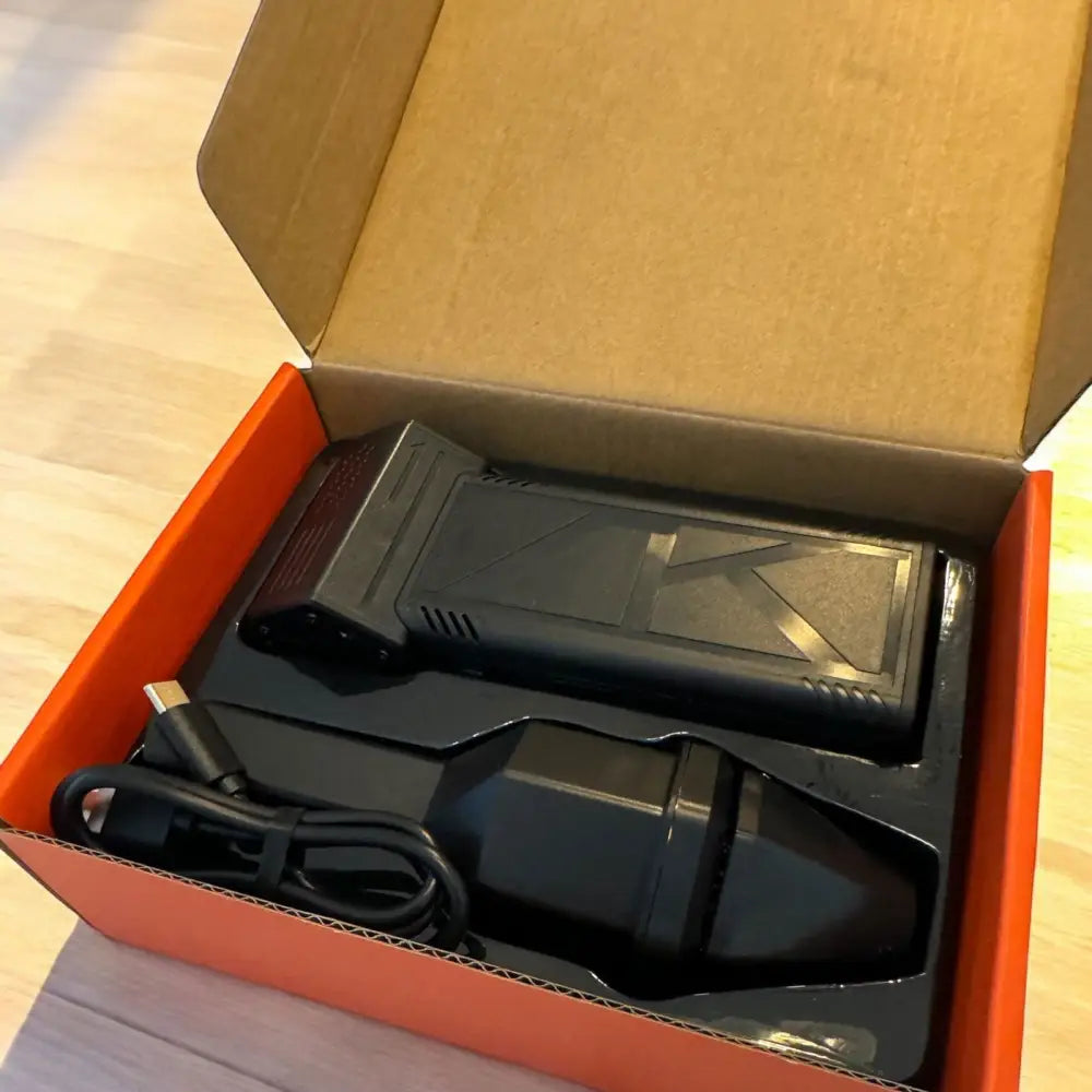 Black plastic drone battery with textured surface and angular design, nestled in a molded black foam insert inside an orange cardboard box.