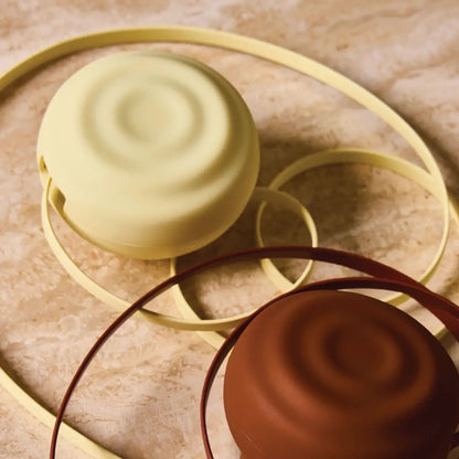 Two round, minimalist handbags with smooth, matte-finished silicone bodies in pale yellow and terracotta brown, each featuring a matching colored loop handle.